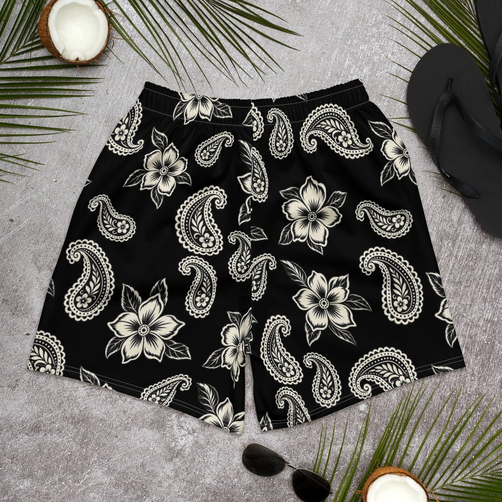 Black athletic shorts with cream floral and paisley print, displayed with palm leaves and sunglasses.