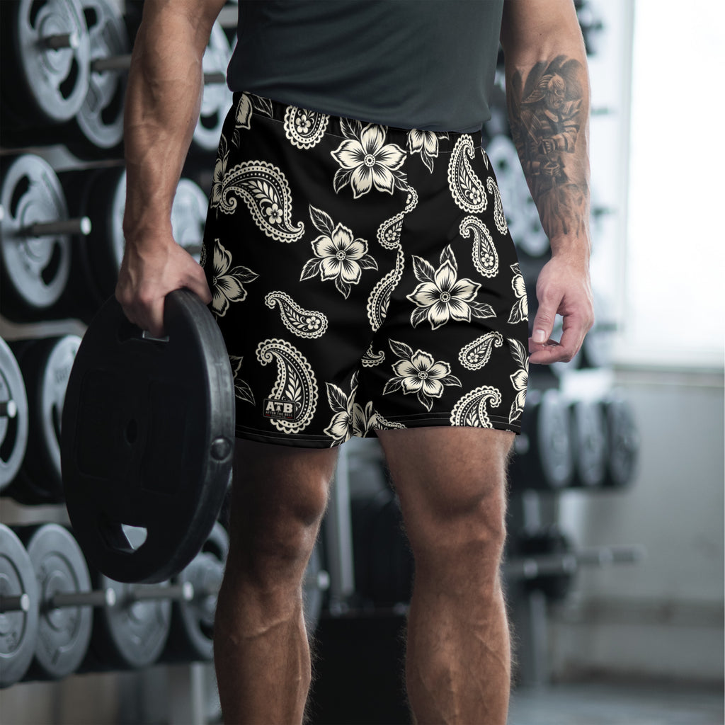 Man wearing black floral and paisley athletic shorts while holding a weight plate in a gym.