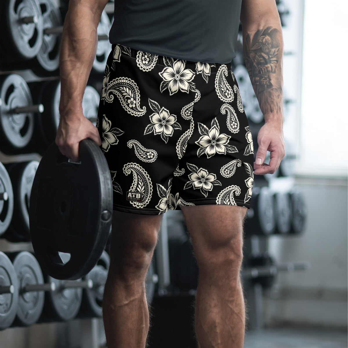Man wearing black floral and paisley athletic shorts while holding a weight plate in a gym.